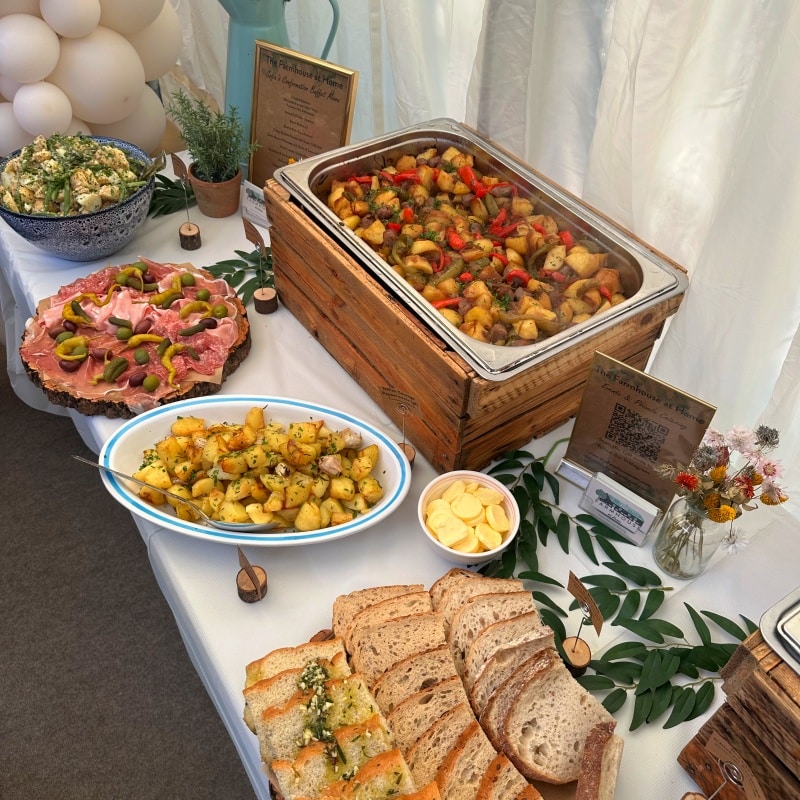 A buffet table featuring various dishes including a tray of roasted vegetables, a platter of roasted potatos, and a dish of cured meat, along with small potted plants and decor.